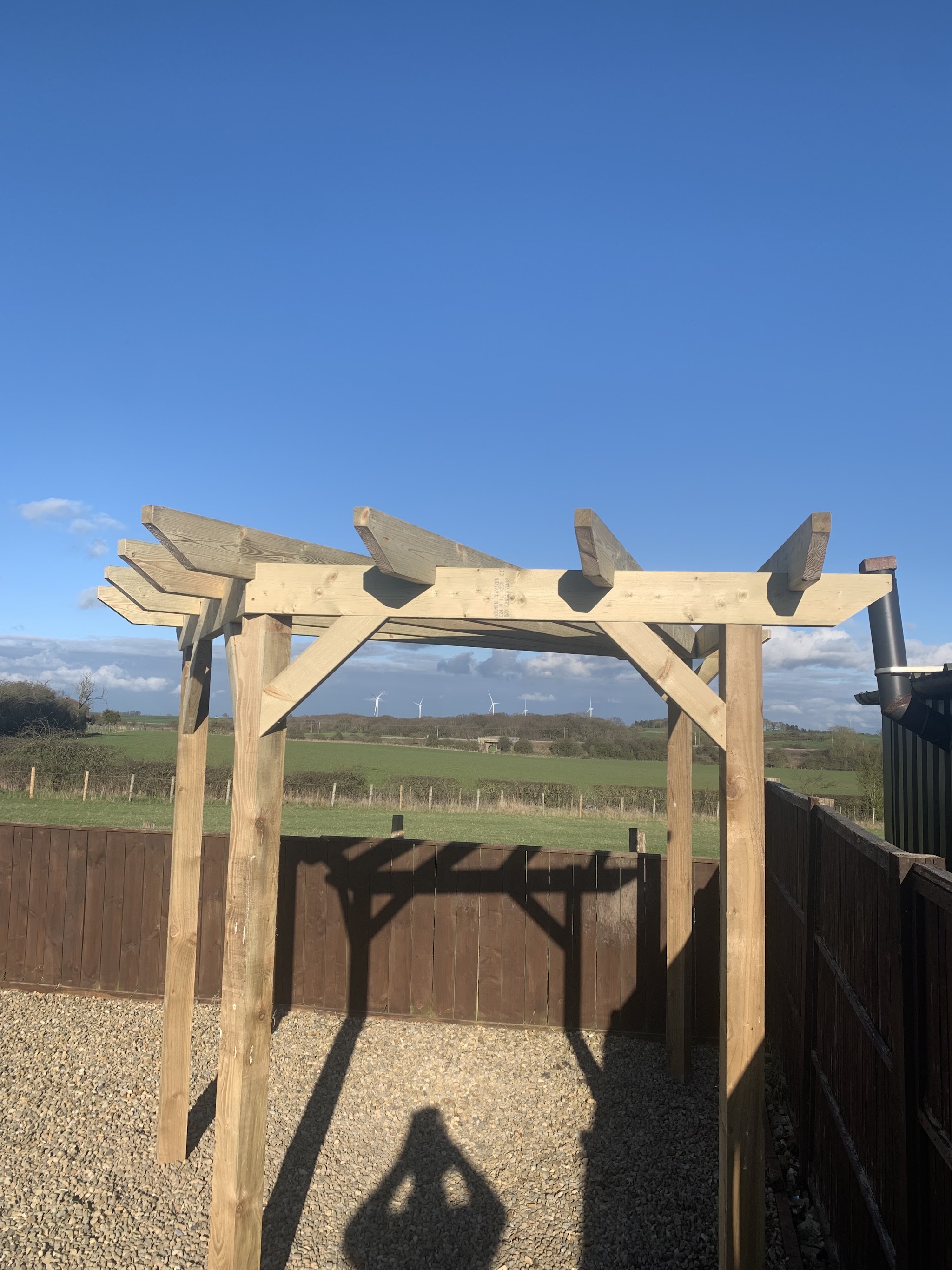 New timber pergola standing on gravel under a clear blue sky, with fields beyond the fence